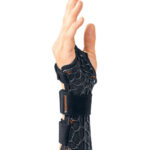 Dłoń w opasce ortopedycznej z czarnej siateczki, uniesiona w górę, z palcami lekko rozłożonymi.