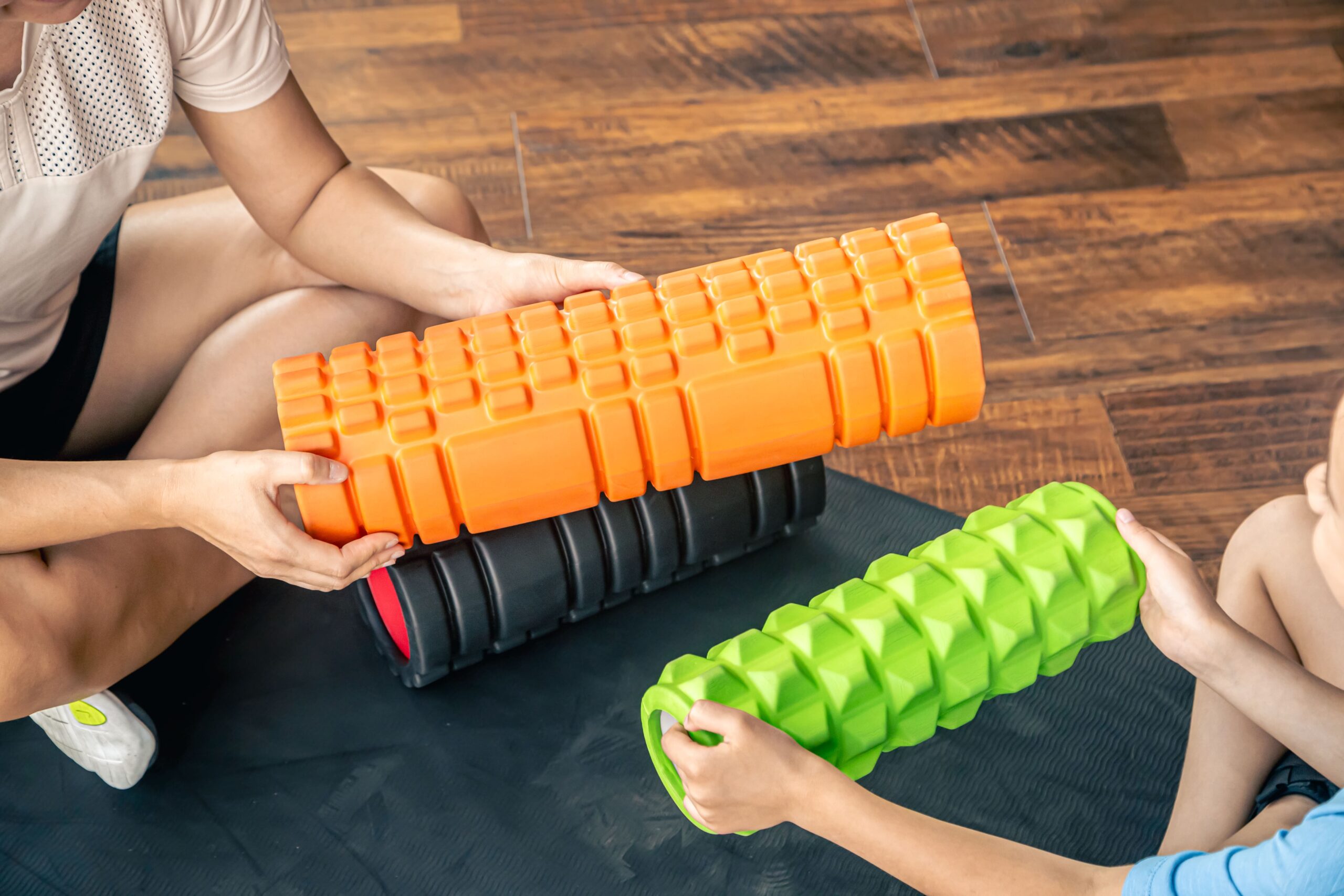 Dwie osoby trzymają kolorowe wałki do masażu o teksturowanej powierzchni, siedząc na matce na drewnianej podłodze.