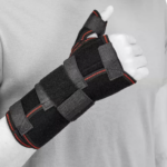 Osoba nosi czarny stabilizator nadgarstka z czerwonymi akcentami, obejmujący dłoń i nadgarstek, z zamkiem na kciuku.