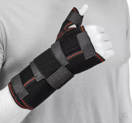 Osoba nosi czarny stabilizator nadgarstka z czerwonymi akcentami, obejmujący dłoń i nadgarstek, z zamkiem na kciuku.