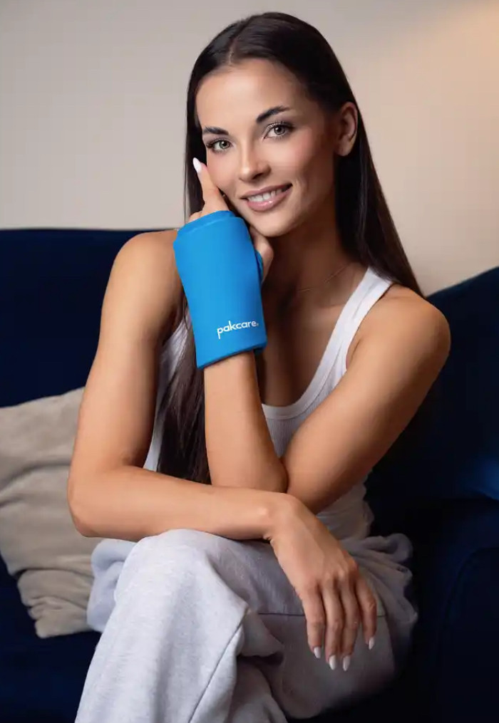 Uśmiechnięta kobieta z długimi brązowymi włosami, nosząca białą koszulkę i szare spodnie, trzyma rękę w specjalnym